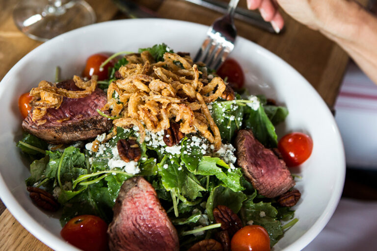 steak salad served in a bowl with tomatoes, friend onions and blue cheese crumblings